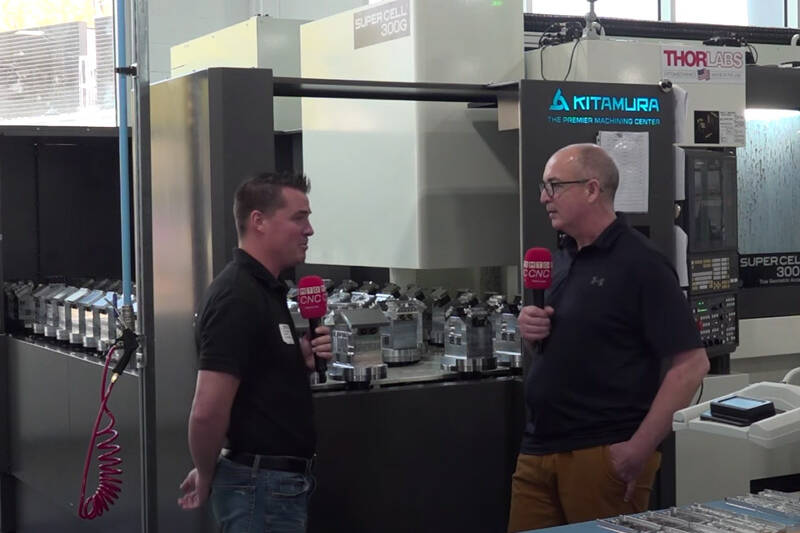 Two people talking into microphones next to large machines.