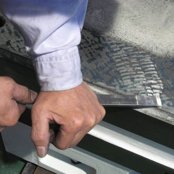 A person uses a hand scraper tool to remove damaged material from a surface, wearing a light blue long-sleeve shirt.