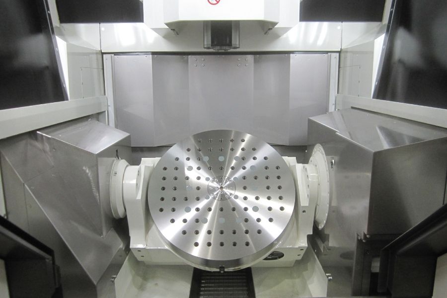 Interior view of a 5-axis CNC machining center featuring a large rotary table with multiple fixture holes for high-precision part positioning.
