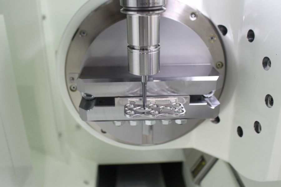 Close-up of a 5-axis CNC machine performing precision milling on a small, complex metal component held in a custom fixture on a rotary table.