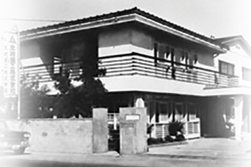 Black-and-white photo of Kitamura Machinery's original 1960s two-story headquarters with a tiled roof and signage in Japanese.