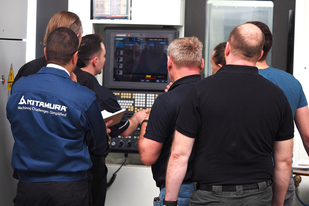 Multiple people gathered around a Kitamura CNC control panel during a hands-on training session, viewing the display screen.
