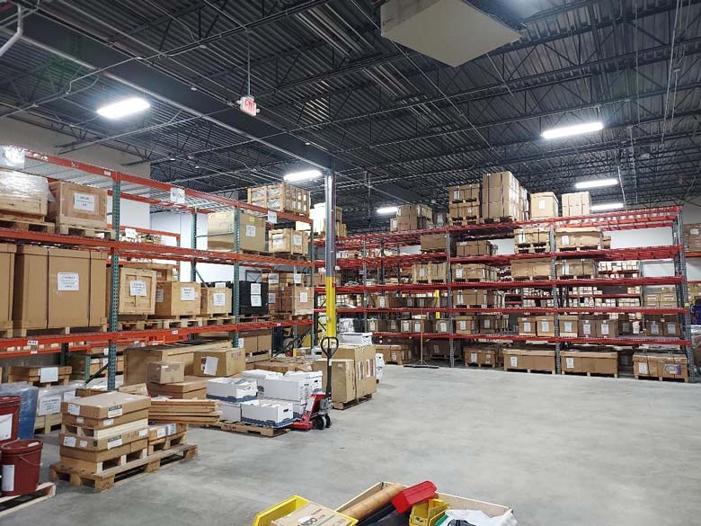 **Alt text:** Warehouse interior with industrial shelving stacked with labeled boxes and pallets, under bright overhead lighting.